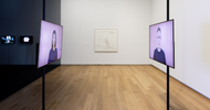 two large video monitors show two people facing each other with neutral expressions. They are infront of lilac backgrounds. In the background there is a white gallery space with a large drawing on the backwall, on the left side are two small video monitors.