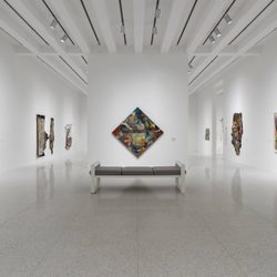 A modern art gallery with white walls, a bench in the center, and various colorful abstract paintings displayed on the surrounding walls. The space is brightly lit with overhead lights.
