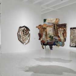 Contemporary art gallery with mixed-media abstract artworks on white walls and a large, textured sculpture suspended in the center. The gallery has polished floors and high ceilings.