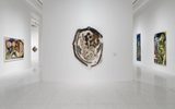 A modern art gallery with white walls displays abstract paintings and textile art. One large textile artwork hangs on the central wall, surrounded by colorful abstract paintings on adjacent walls.