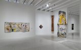A modern art gallery with abstract paintings on white walls and a tall, colorful mixed-media sculpture hanging from the ceiling. The room is bright with spotlights illuminating the artworks.