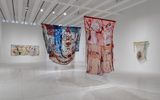 A modern art gallery with three large, colorful, abstract textile artworks hanging from the ceiling and one abstract painting displayed on the white wall in the background.