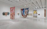 Contemporary art gallery with colorful abstract textile and mixed-media artworks hanging on white walls and suspended from the ceiling under bright lights.