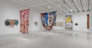Contemporary art gallery with colorful abstract textile and mixed-media artworks hanging on white walls and suspended from the ceiling under bright lights.