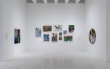 A white gallery room displays colorful abstract paintings on three walls, including a cluster of nine rectangular pieces and a single round portrait, all under bright lighting.