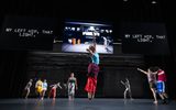 A group of dancers perform on a dimly lit stage; one leaps high in the center. Behind them, large screens display the text MY LEFT HIP, THAT LIGHT. and a black-and-white photo of an industrial space.