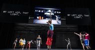 A group of dancers perform on a dimly lit stage; one leaps high in the center. Behind them, large screens display the text MY LEFT HIP, THAT LIGHT. and a black-and-white photo of an industrial space.