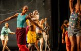 A group of diverse dancers performs on stage, each striking expressive poses. They wear colorful, varied outfits and move energetically in front of a dark, minimalist backdrop.