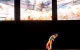 A person in orange pants and a blue shirt poses with arms stretched to the side on a dark stage, beneath three large, abstract, colorful screens.