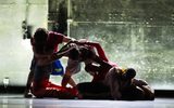 Five dancers in colorful costumes perform a contemporary routine, intertwining their bodies in a low, expressive formation on a dimly lit stage with a textured, metallic background.