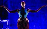 A dancer in a blue tank top and maroon pants stands with arms outstretched against a dark blue background, while another persons hands support their waist from behind.