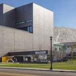 A person walking on a sidewalk in front of the entrance of a gray modern-shaped building, the Walker Art Center.