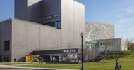 A person walking on a sidewalk in front of the entrance of a gray modern-shaped building, the Walker Art Center.