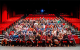 A large theatre with red seats and black stair railings, with mostly the middle section filled with smiling adults posing for a group photo