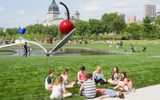Groups of people enjoying a park with a large outdoor sculpture of a cherry atop a spoon, floating over a small man-made lake, surrounded by grass, trees, and buildings