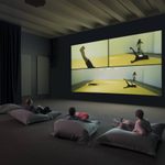 Three people lying on large pillows on the ground, watching a wall-sized screen of another person on a yellow mat doing movements