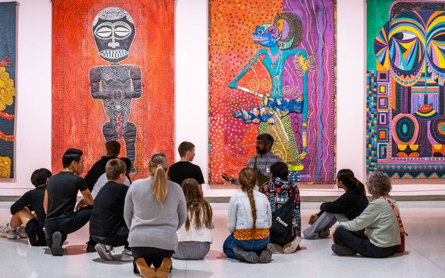 A dozen patrons sit on the floor while listening to a person speak in front of large colorful cultural artworks hanging on the wall
