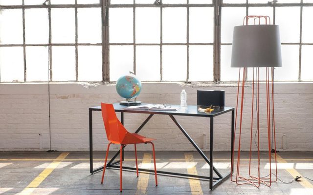 A black framed desk with a blue globe, white book open with eyeglasses lying ontop, next to a banana peel and water bottle. And an orange chair and tall grey and orange lamp next to large warehouse windows with sun shining in