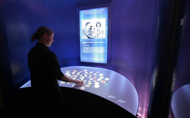 An adult with their hair tied back touches a digital display that shows digital historical slides on a screen above the display.