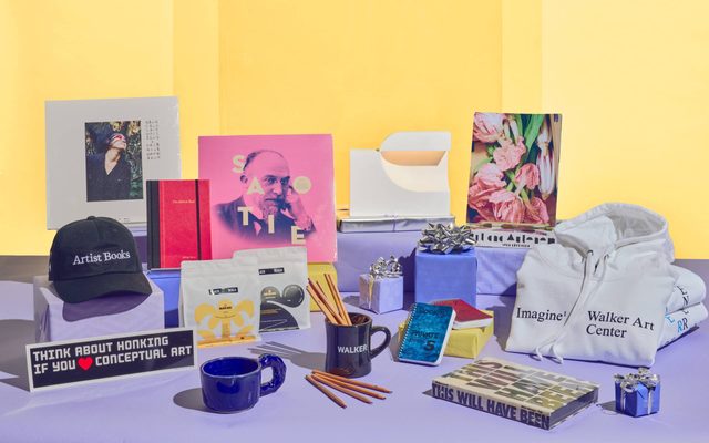 Display of Walker Art Center merchandise, including a hat, mugs, pencils, sweatshirt, notepads, books, sticjers and tiny gift boxes with silver bows.