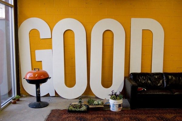 Large cutout letters spelling GOOD lean against an orange wall in front of a BBQ grill and black leather couch, with a red rug and large window.