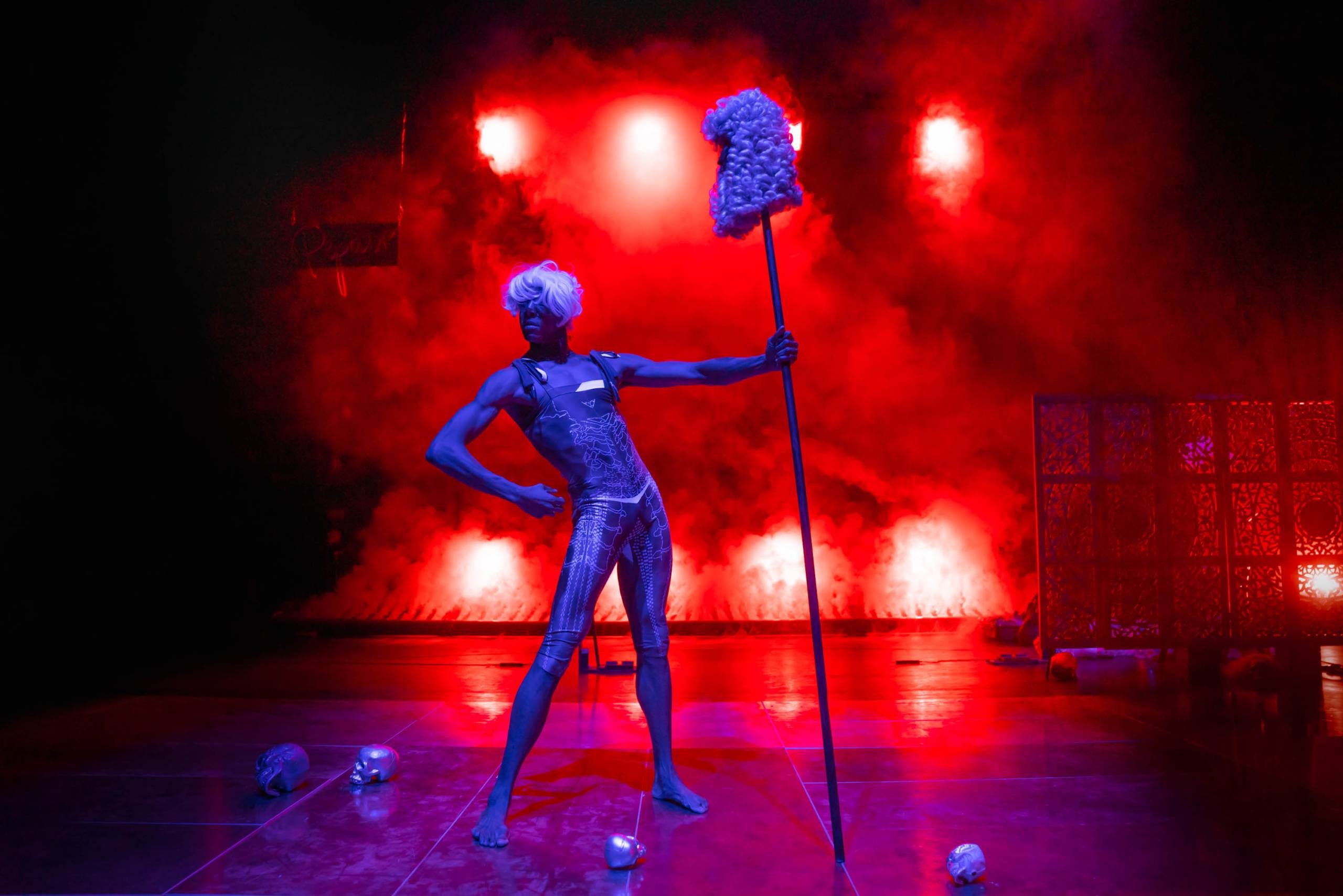 A live artist on stage holding a stick with a parliament-style wig on top, with small silver skulls on the ground in front of smoke and red lights