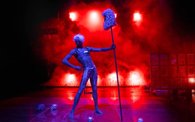 A live artist on stage holding a stick with a parliament-style wig on top, with small silver skulls on the ground in front of smoke and red lights
