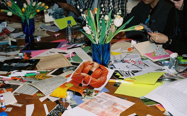 A cluttered table with papers, glue, flowers, and people crafting and cutting out magazine images.