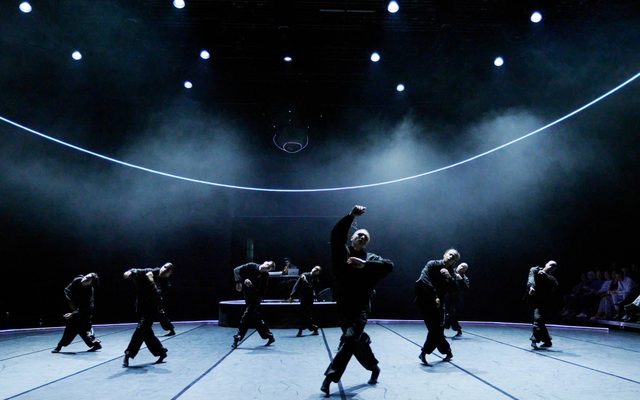 A hooded dancer crosses their legs with arms outstretched.