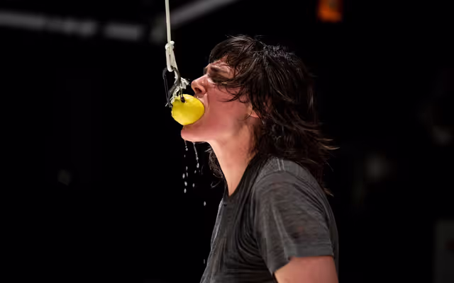 Person with long dark hair bites into a lemon hanging on a hook and rope and lemon juice drips from it