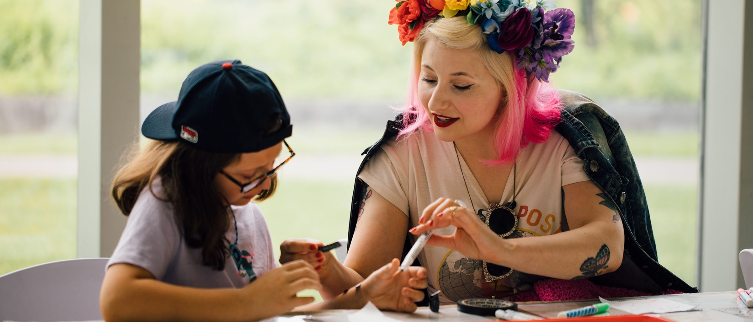 An adult with blonde and pink hair and a flower crown is sitting at a table working on art with a child wearing a backwards baseball cap