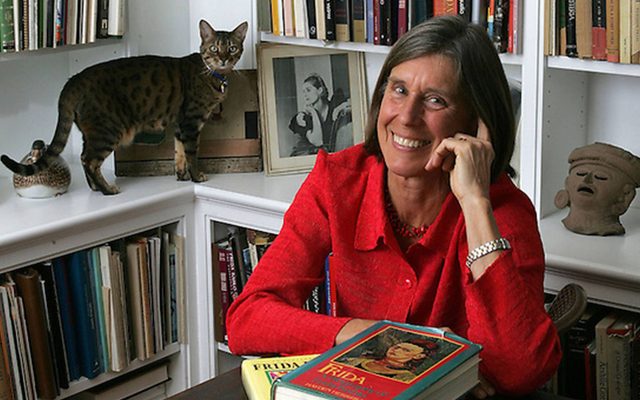 An adult with light skin, wearing a red shirt and silver watch, has medium legth brown hair and smiles at the viewer with a hand to their face. A cat stands on a bookshelf behind.