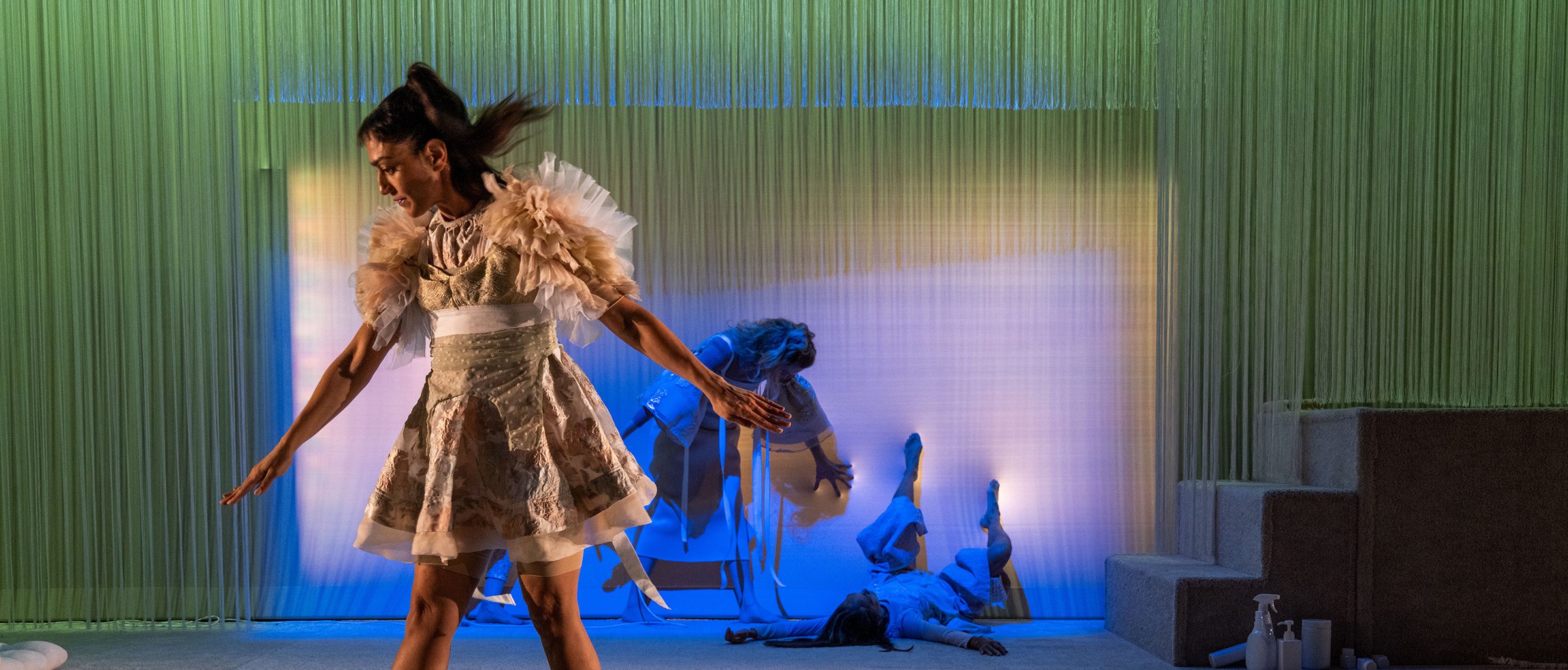 Adult in a elaborate dress posed within a live art installation, with green drapery, small steps, and other adults laying within the installation.