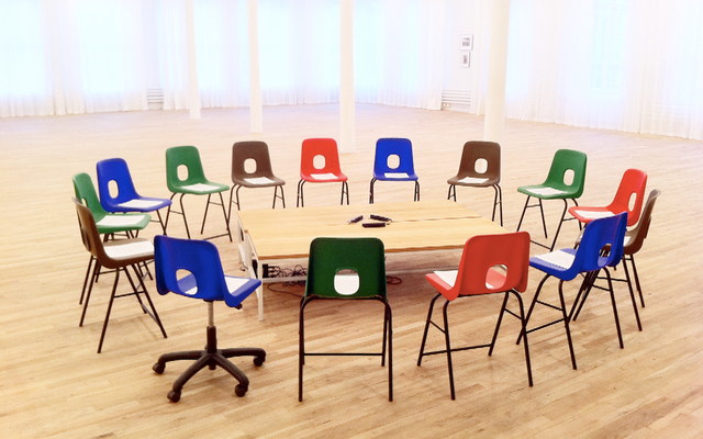 a circle of multicolor chairs facing inwards towards a short square platform in a large lit room with wood floors.