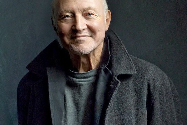 A short haired adult with a black colored coat and grey shirt smiling and posing for a photo
