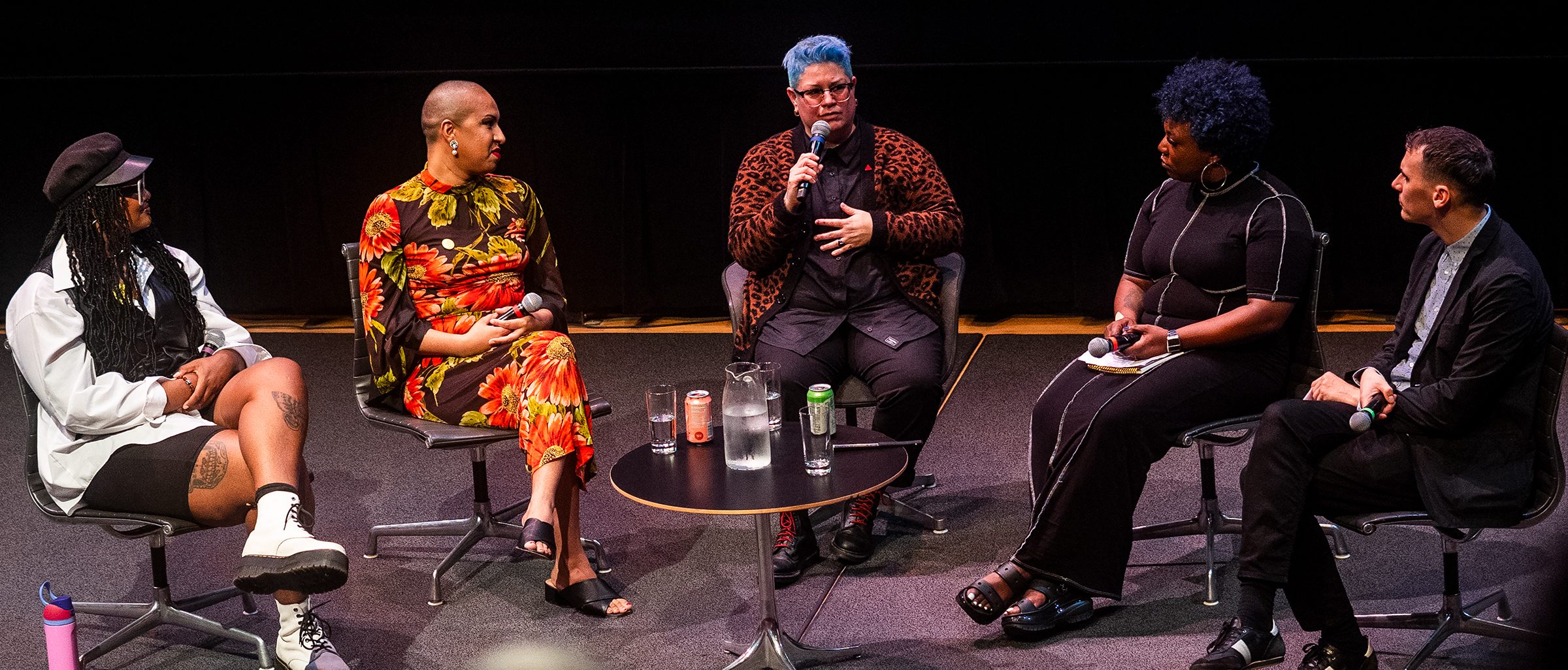 5 adults sit around a small round coffee table with drinks and one person with blue hair is speaking on a microphone while others listen