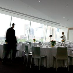Inside of a white dining area with big windows, chairs, tables, glassware, centerpieces, and one staff member