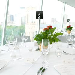 Round dining tables with white table cloth bearing silverware, wine glasses, plant center piece, and a black table sign reading 