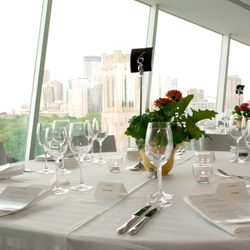 Round dining tables with white table cloth bearing silverware, wine glasses, plant center piece, and a black table sign reading 