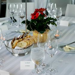 Round dining table with white table cloth bearing silverware, wine glasses, plant center piece, bread, butter and tealight candles