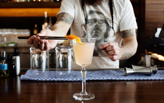A bartender with a white T-shirt places a curled orange peel pink-colored drink in a long stem glass with chopsticks, at a bar top with blue cloth and bar items.