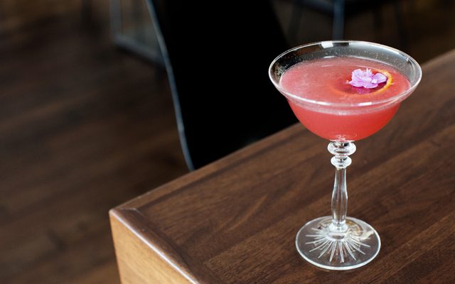 A long-stemmed glass drink with red fluid and a purple flower atop a brown table next to a black chair