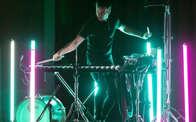 One person with light skin and short hair plays a xylophone with blue and pink vertical fluorescent lights standing next to them.