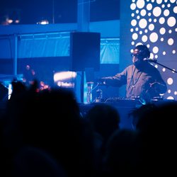 A DJ with a mask and eyeglasses plays to a crowd of people in a blue lit room with a white dot backdrop