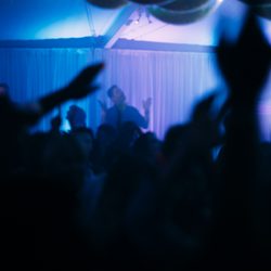 A blue and purple light room with a DJ working a crowd of party goers with their arms in the air