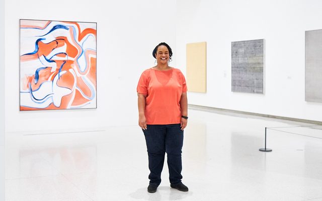 An adult with a coral shirt and dark pants, poses and smiles for a photo in front of displayed artwork in a museum