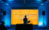 A DJ stands at a table with his gear in front of an orange screen reading 