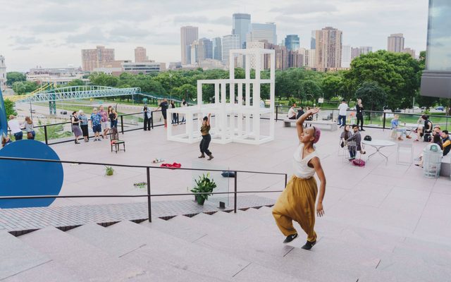 An adult with brown skin, wearing yellow pants and a flowy white top is mid-pose on stairs, a city view and sculpture and people in the background.