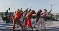 A group of five dancers in colorful, camouflage-inspired outfits strike dynamic poses on an outdoor tiled terrace, with blue sky and cityscape in the background. Music equipment and onlookers are visible behind them.