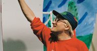 A person wearing glasses, a blue cap, and a red sweatshirt with Walker on it is smiling and reaching upward, with a colorful, abstract background behind them.
